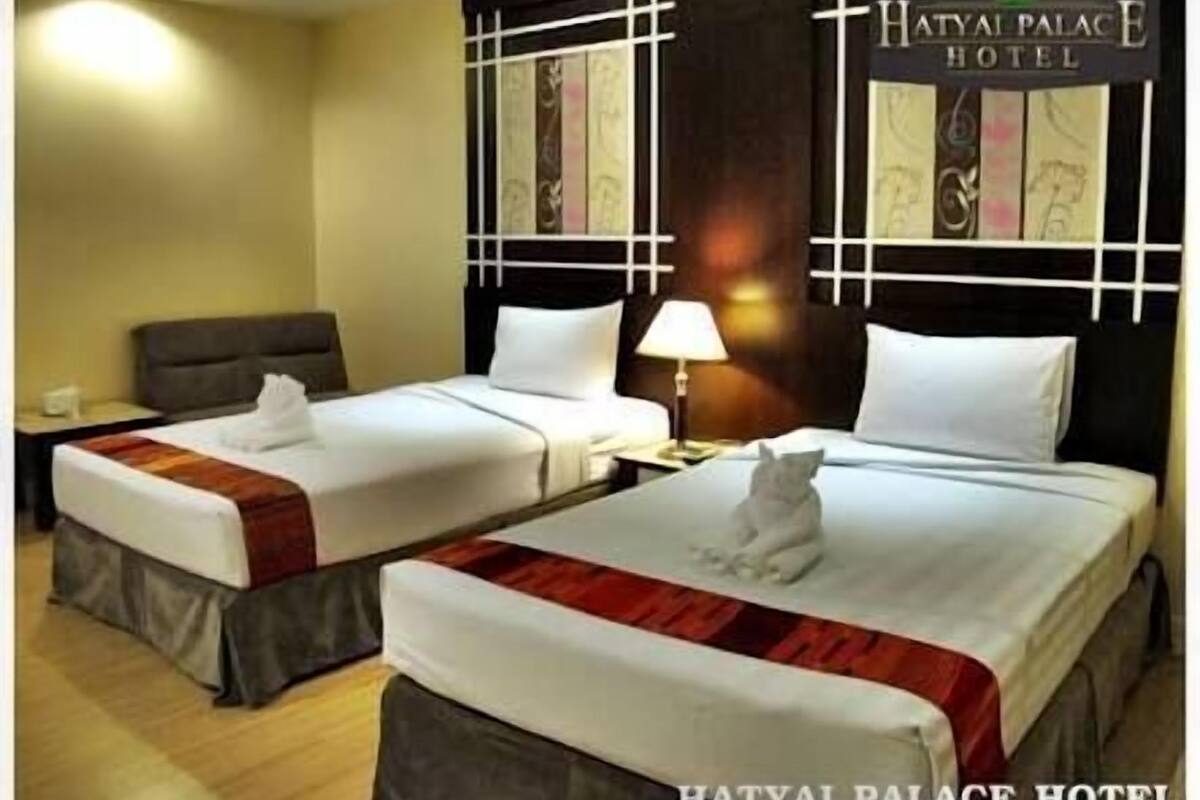 A double room at Hatyai Palace Hotel with two beds, a nightstand, a lamp, and a cat on one bed.