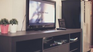A flat-screen TV on a dark wooden entertainment center with a shelf below displaying various electronic devices and books. - Palm Garden Resort (Sikao)
