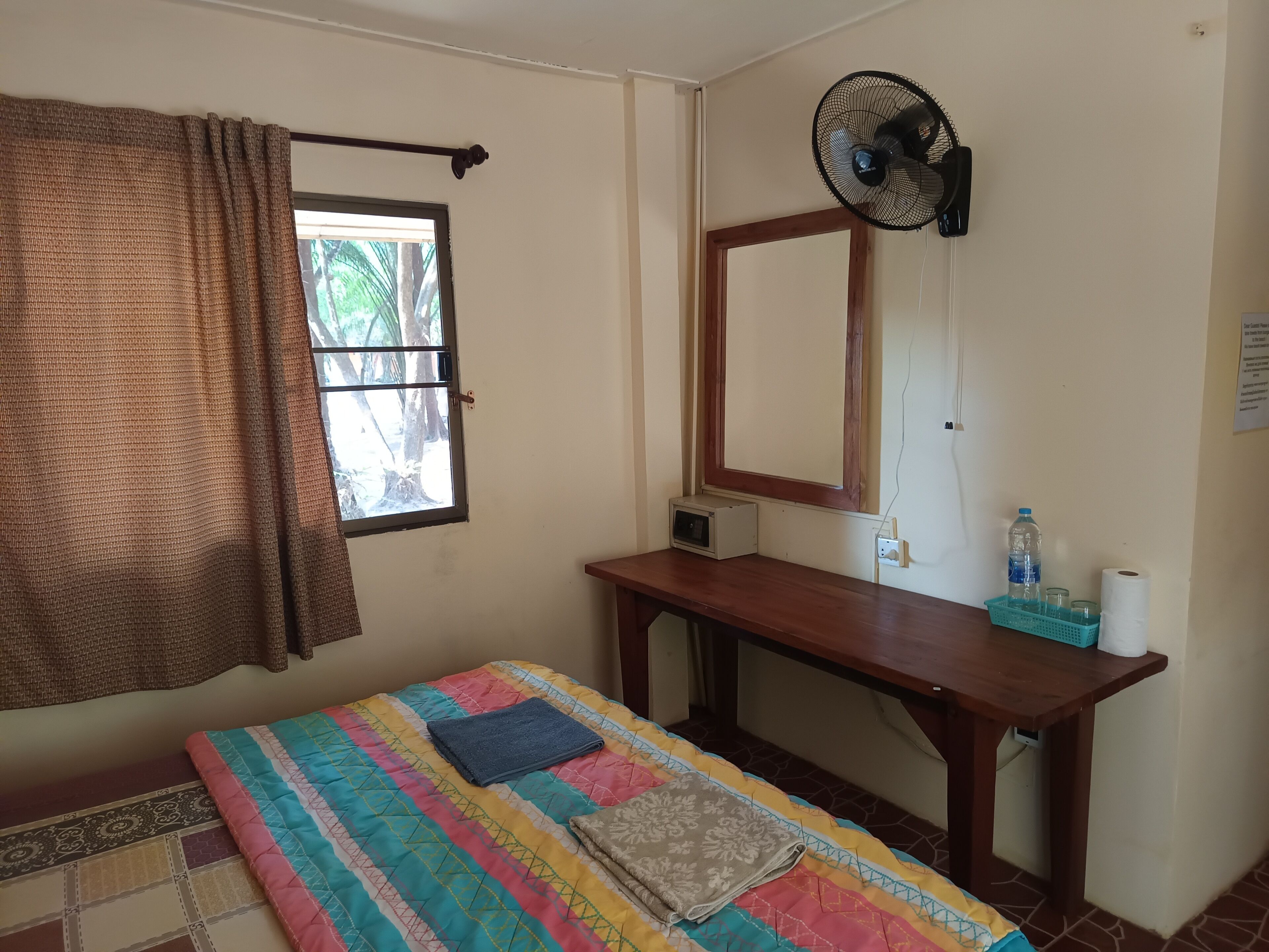 A room with a bed, a desk, a window with a curtain, a wall-mounted fan, and a water bottle on the desk.