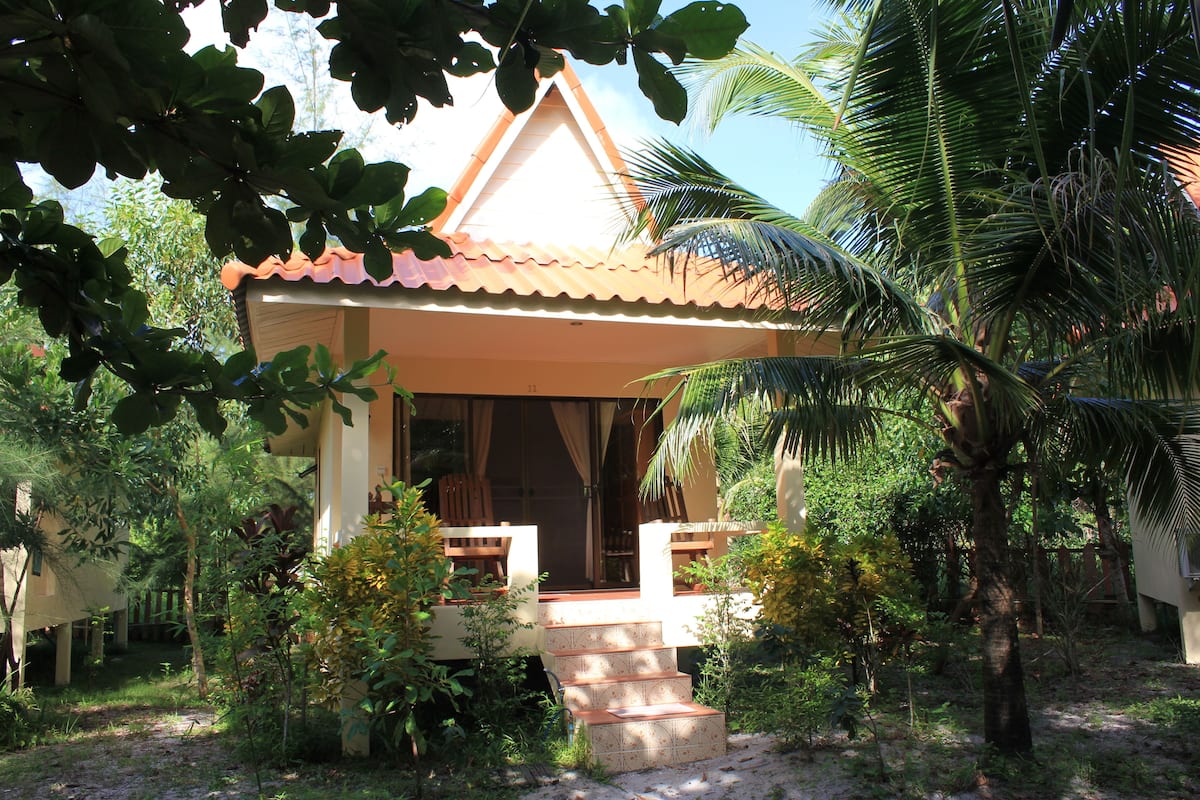 A house with a tiled roof, a porch with steps, and surrounded by greenery including palm trees.