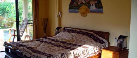 A bedroom with a bed, a nightstand, a wall-mounted fan, and a window with a view of greenery.