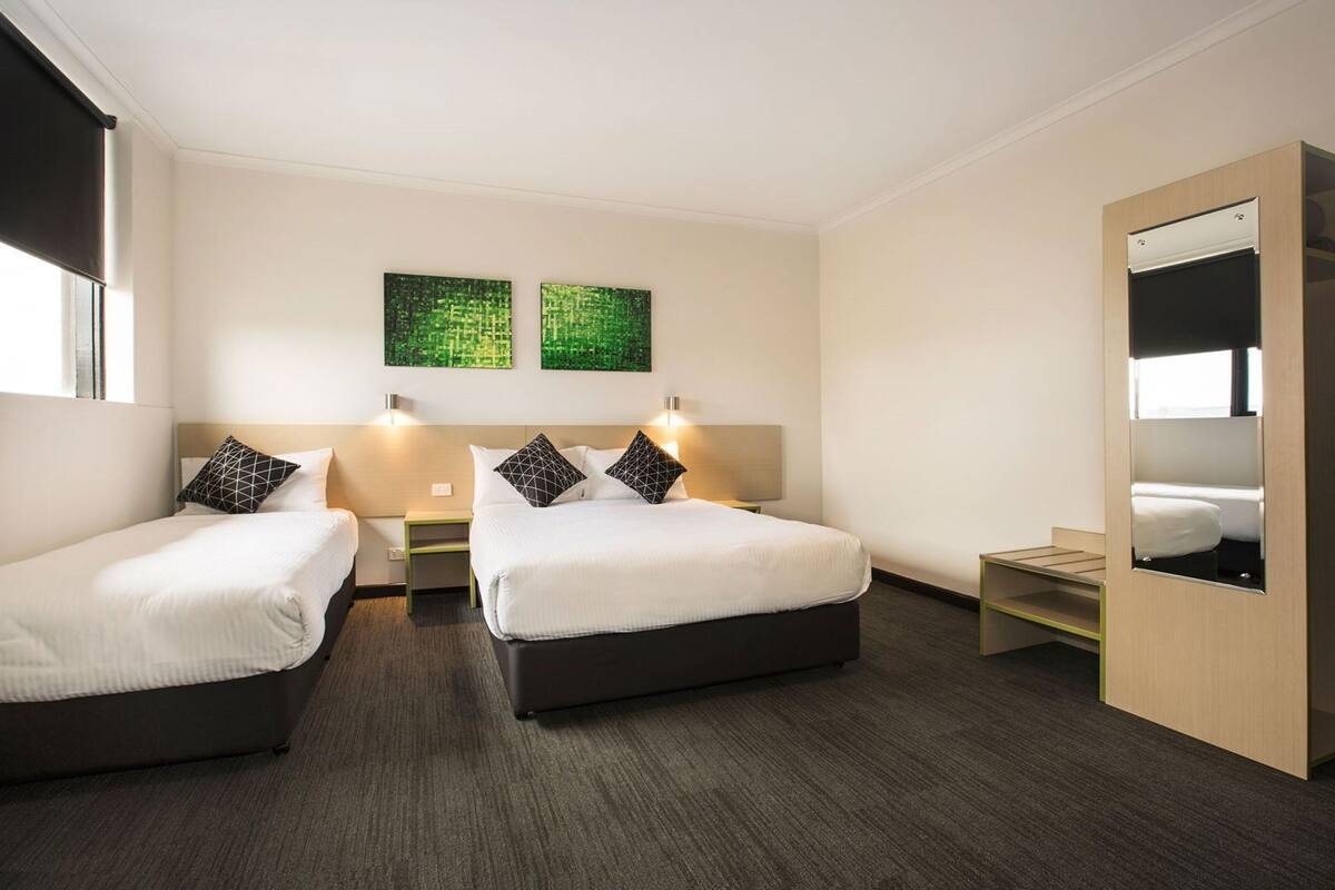 A hotel room with two beds, a small bedside table, a nightstand, a mirror, and two green wall art pieces.
