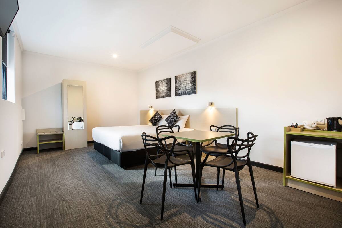 A hotel room with a bed, a desk, a chair, a small table, and a refrigerator.