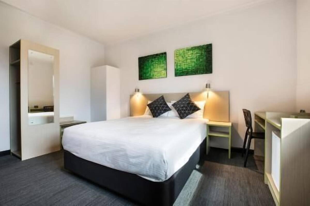 A hotel room with a bed, bedside table, desk, chair, and two wall-mounted artworks.