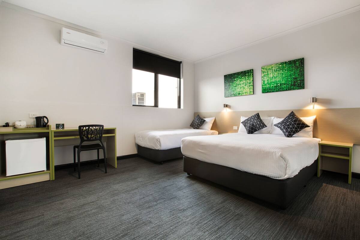A hotel room with two single beds, a desk with a chair, a small kitchenette, and a window with blinds.