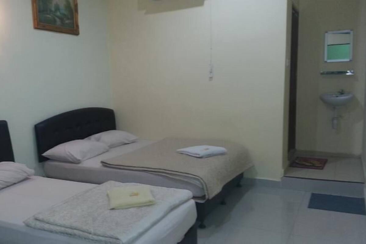 Una habitación pequeña y limpia con dos camas individuales, una mesita de noche, una lámpara de pared y un baño con lavamanos y espejo.