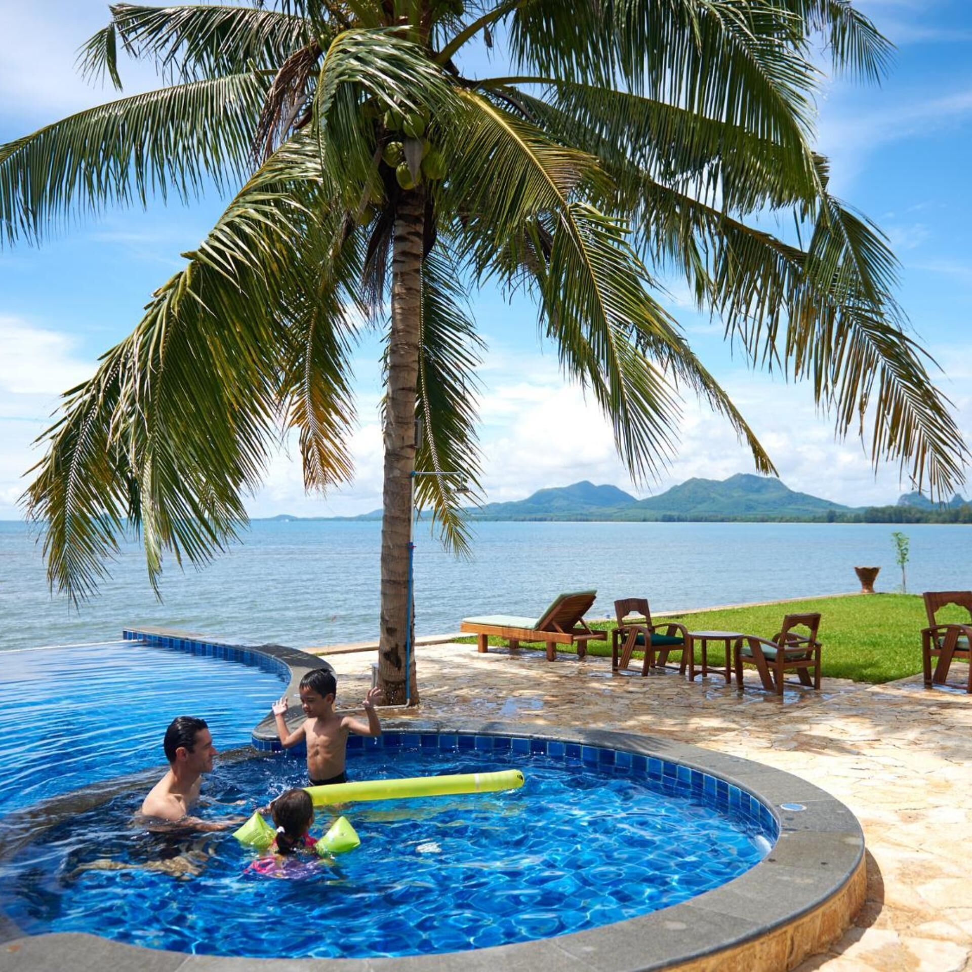 A swimming pool with a hot tub, palm trees, and a view of the sea.