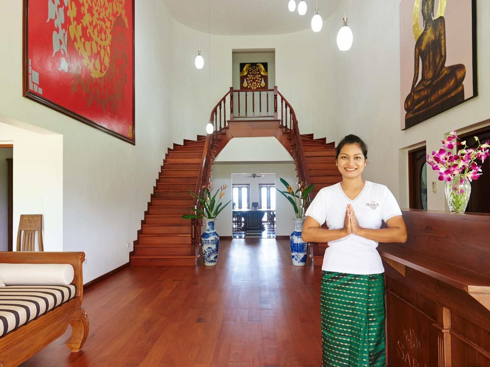 A person standing in a room with a wooden staircase, a large red painting, and a Buddha statue.