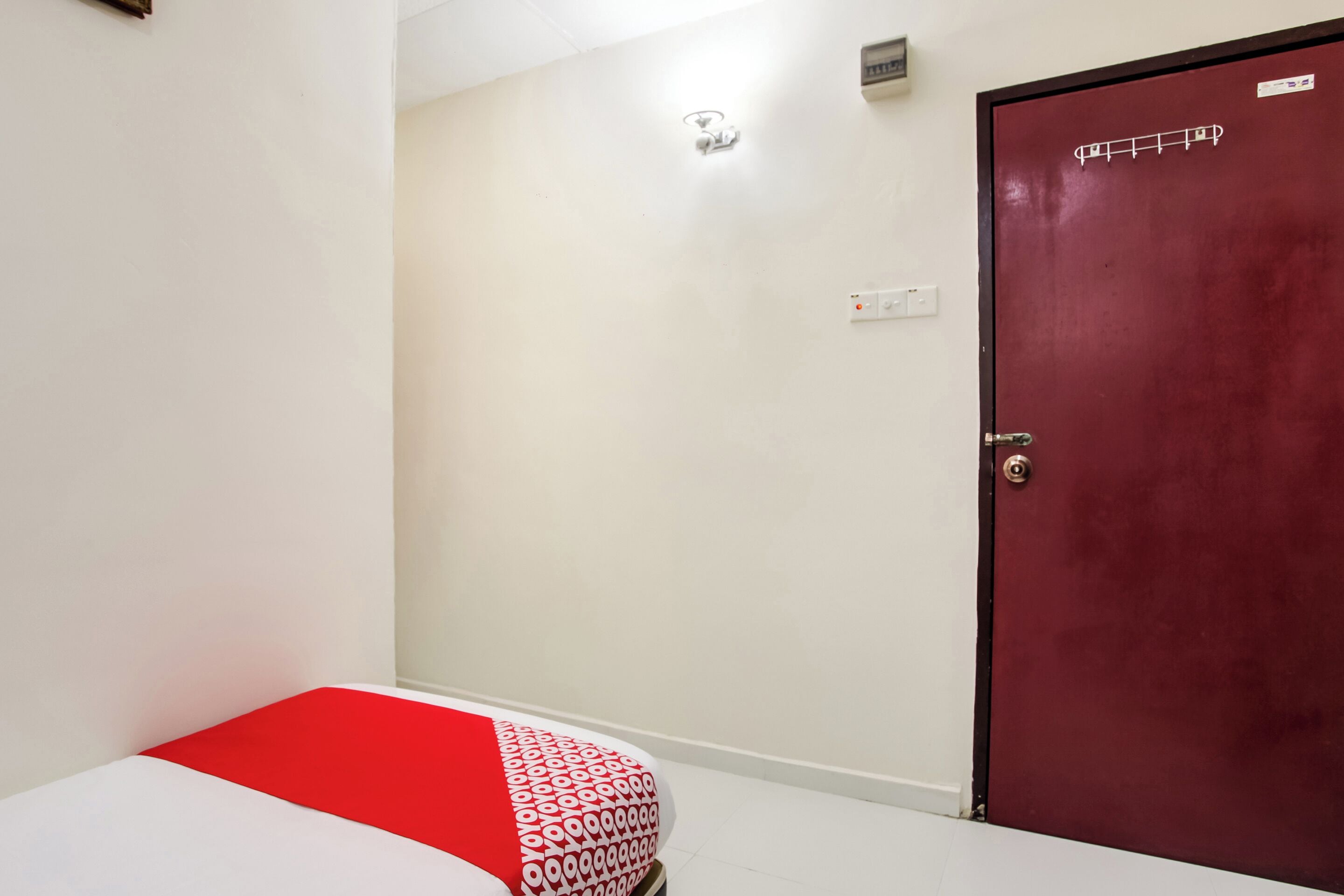 A room with a bed, a red door, and a light switch on the wall.