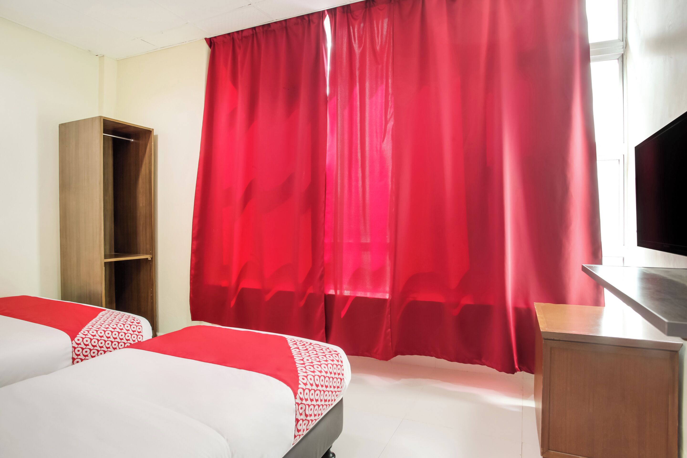 A hotel room with two beds, a wooden wardrobe, a TV, and red curtains.