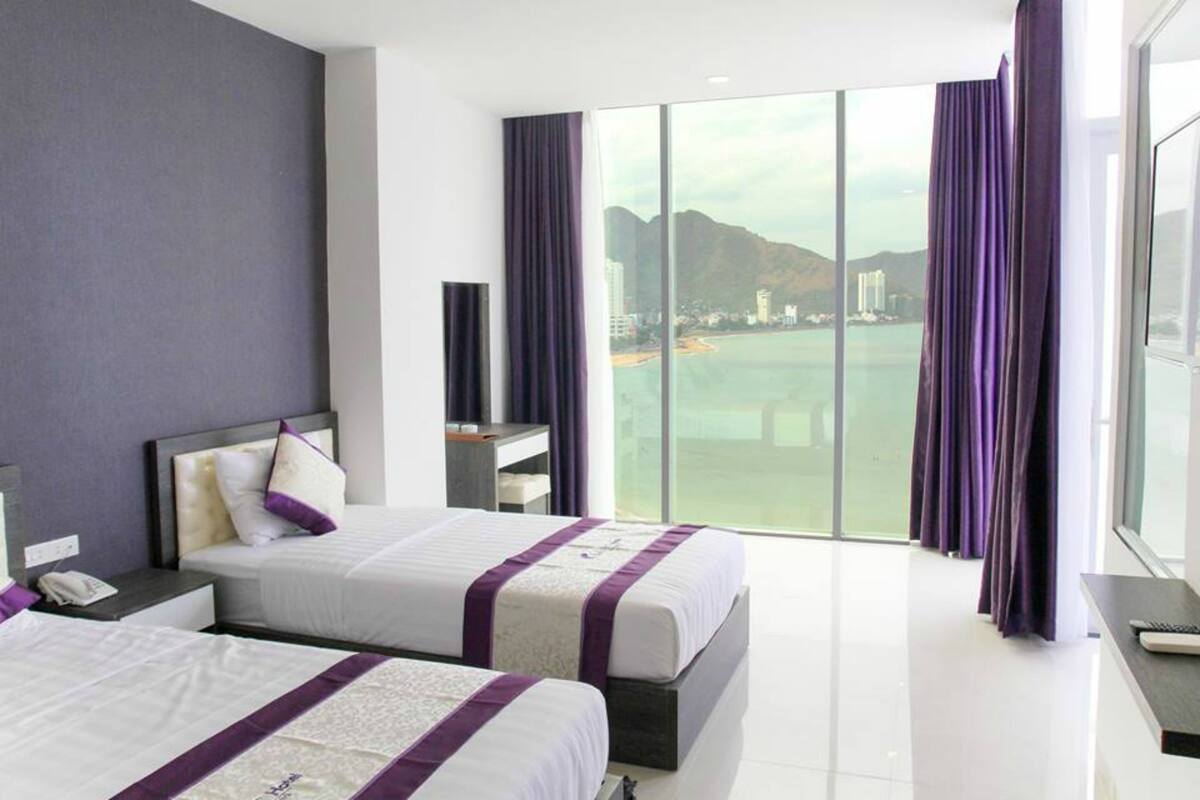 A hotel room with two beds, a large window with a view of a beach and mountains, and purple curtains.