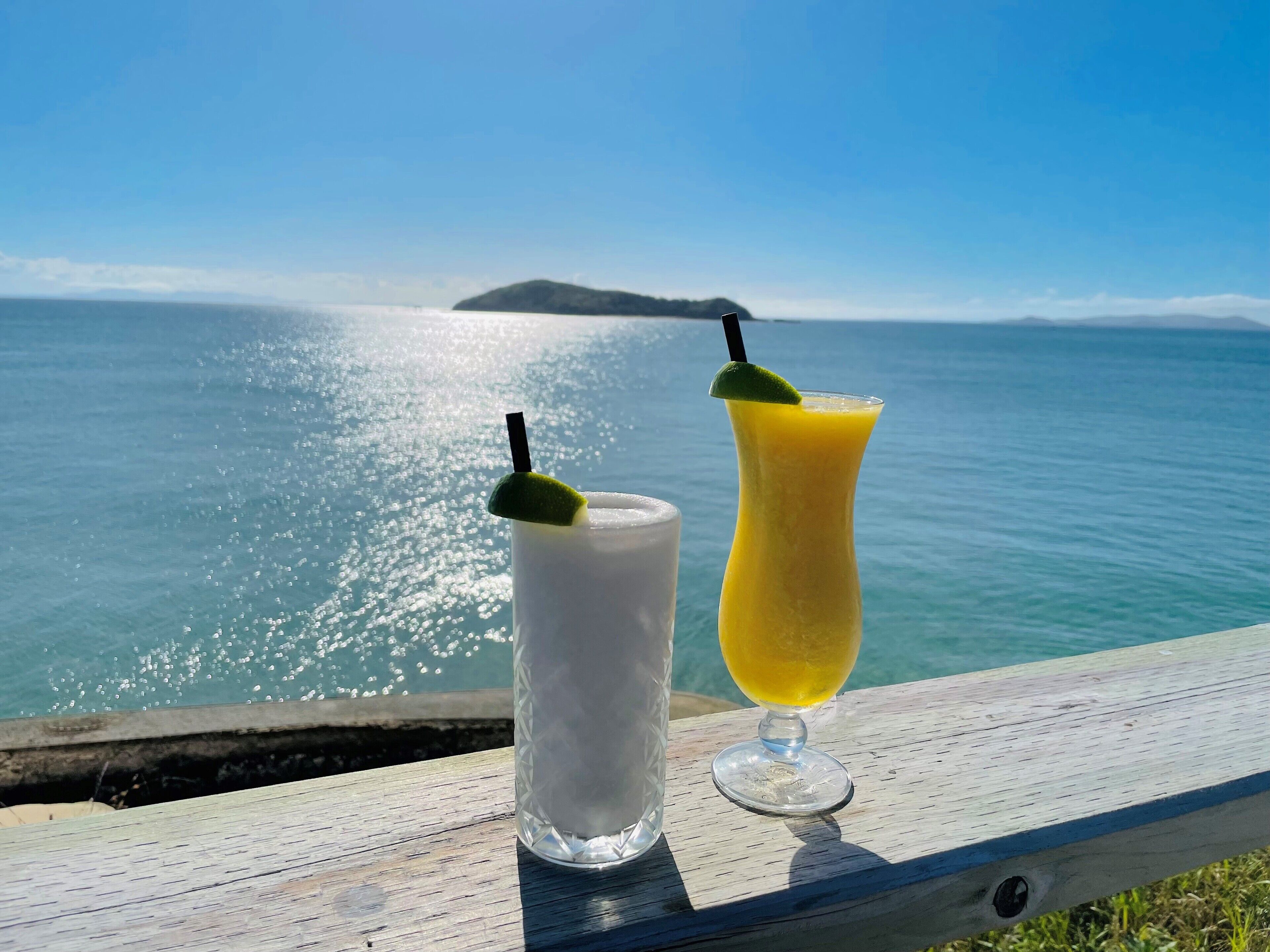 Dos bebidas sobre una terraza de madera con vista al océano y una isla distante.