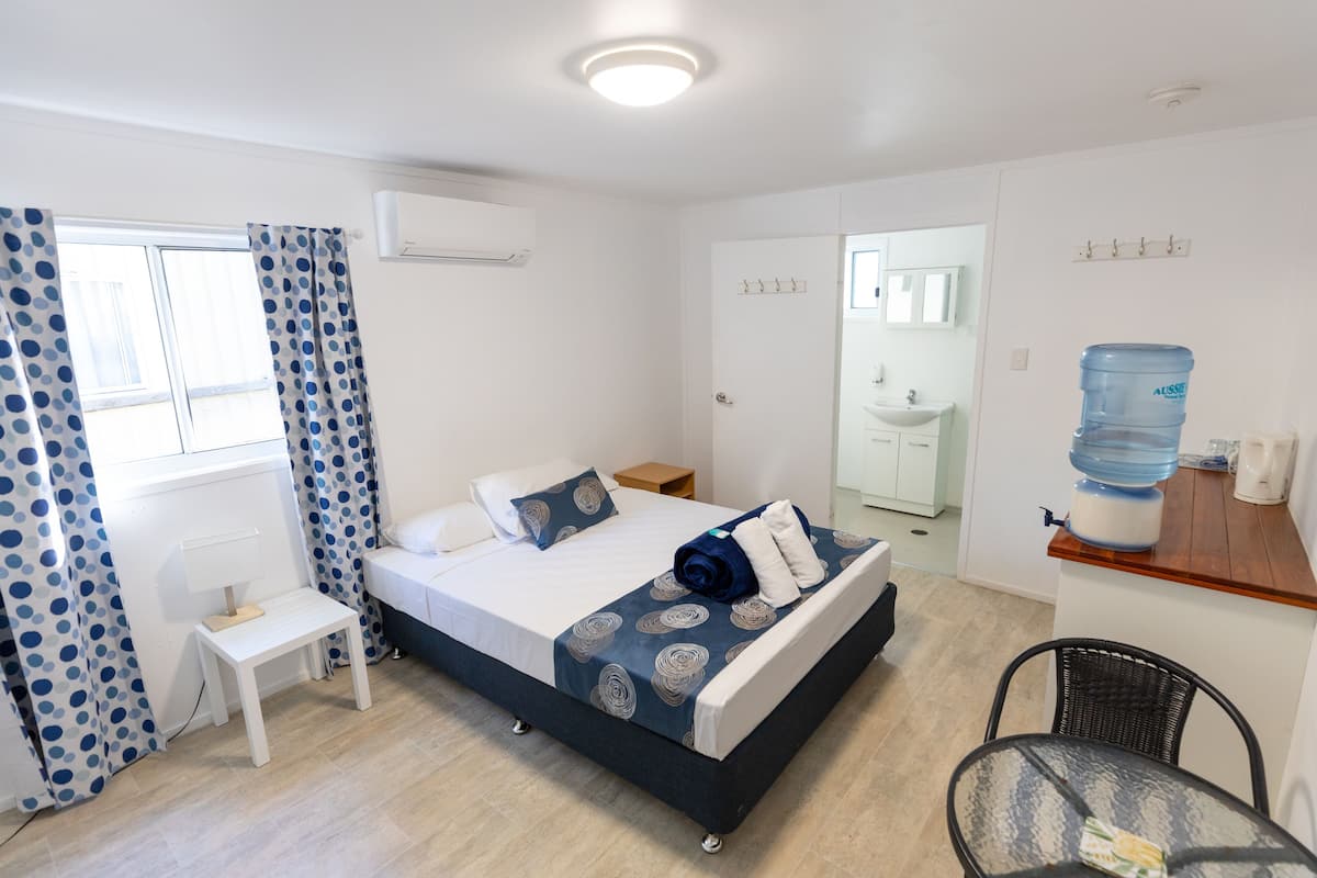 Habitación de hotel con cama, una mesita, una silla, dispensador de agua y baño visible a través de una puerta abierta.