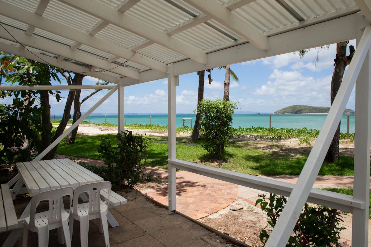 Un espacio al aire libre techado con una mesa y sillas, con vista a una playa con palmeras y el océano.