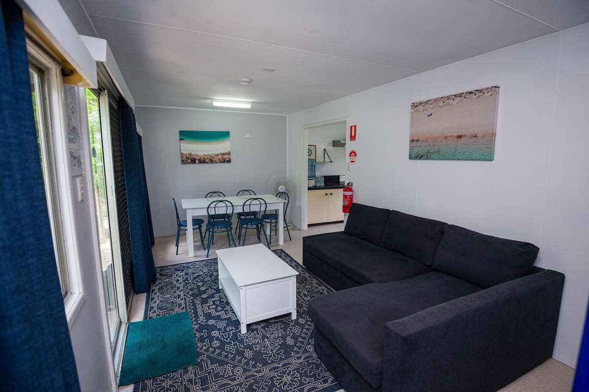 Una sala de estar con un sofá oscuro, una mesa de centro blanca y un tapete estampado. Hay un comedor con mesa y sillas, y una cocina con fregadero y estufa.