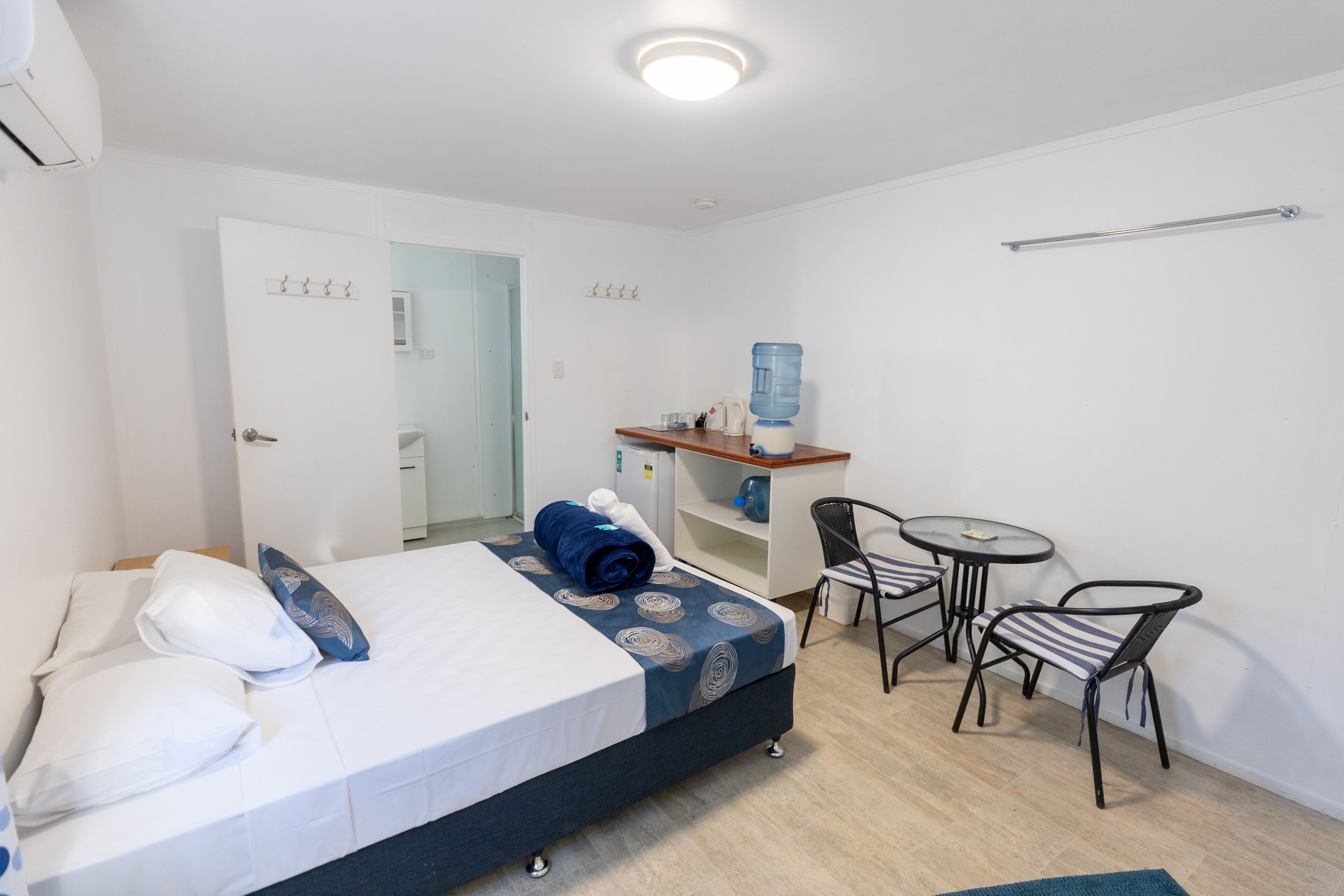 Habitación de hotel con una cama, una pequeña mesa con sillas, un dispensador de agua y un espejo en la pared.