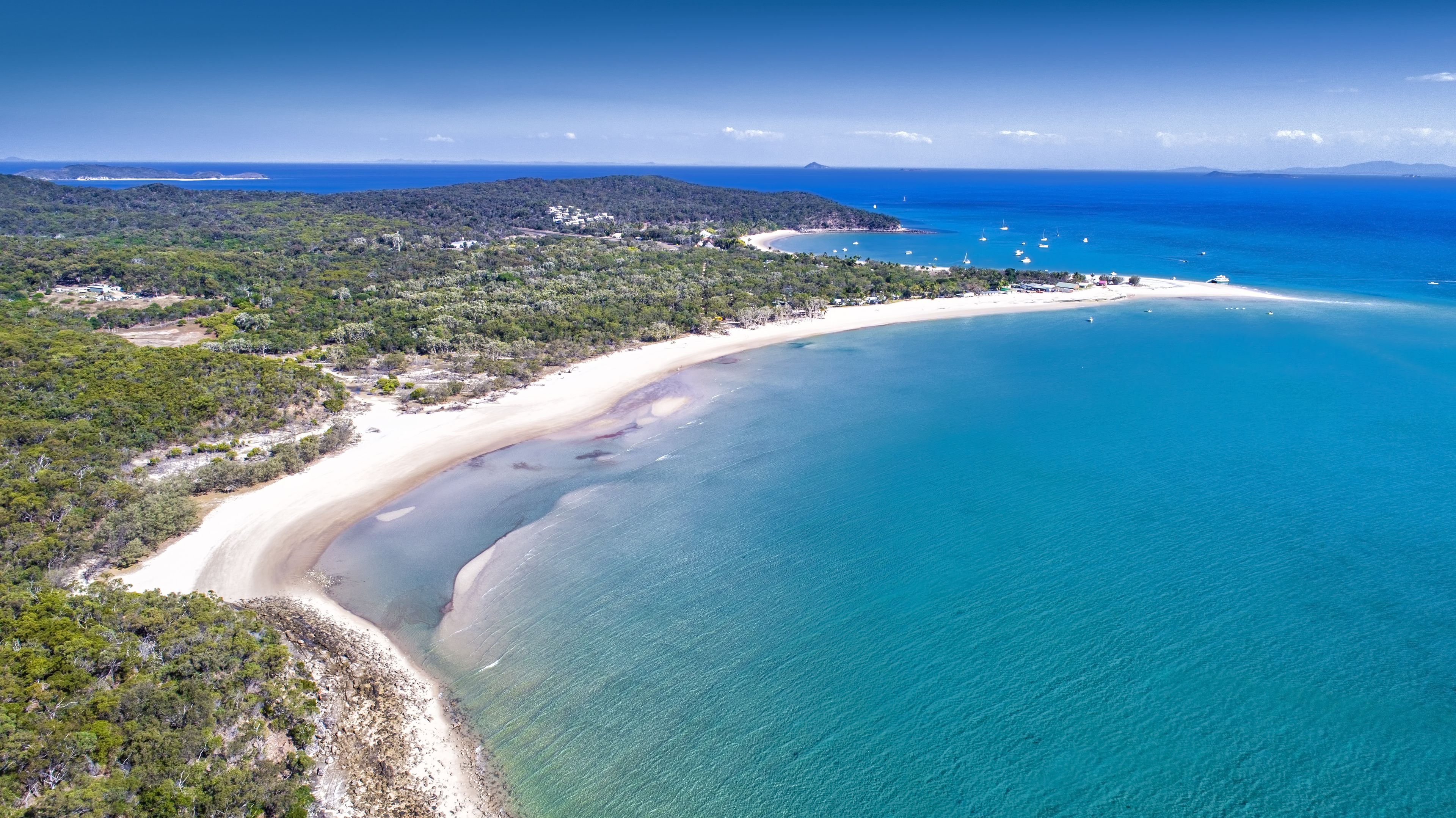 Vue aérienne d’une zone côtière avec une plage de sable, des eaux d’un bleu limpide et, en arrière-plan, des collines et une chaîne de montagnes lointaine.