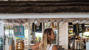 Una mujer está de pie frente a una barra con techo de paja rústico, un fondo de madera blanca y varios objetos, como un tarro de naranjas y un letrero con texto.