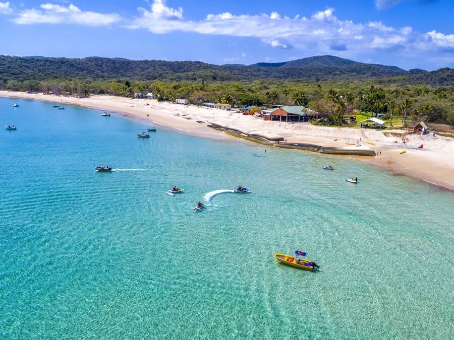 An aerial view of a beach with clear turquoise waters, boats, and a sandy shore with buildings and palm trees.