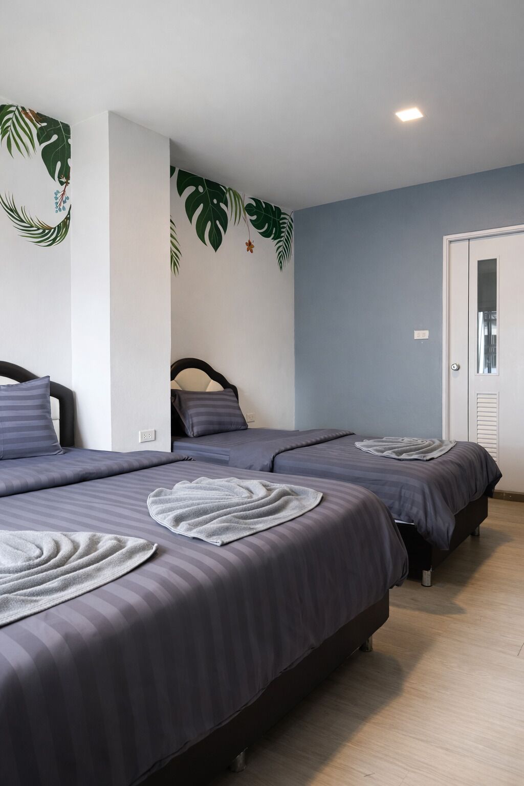 A hotel room with three beds, a white wall with tropical leaf decals, a white door, and a light switch.