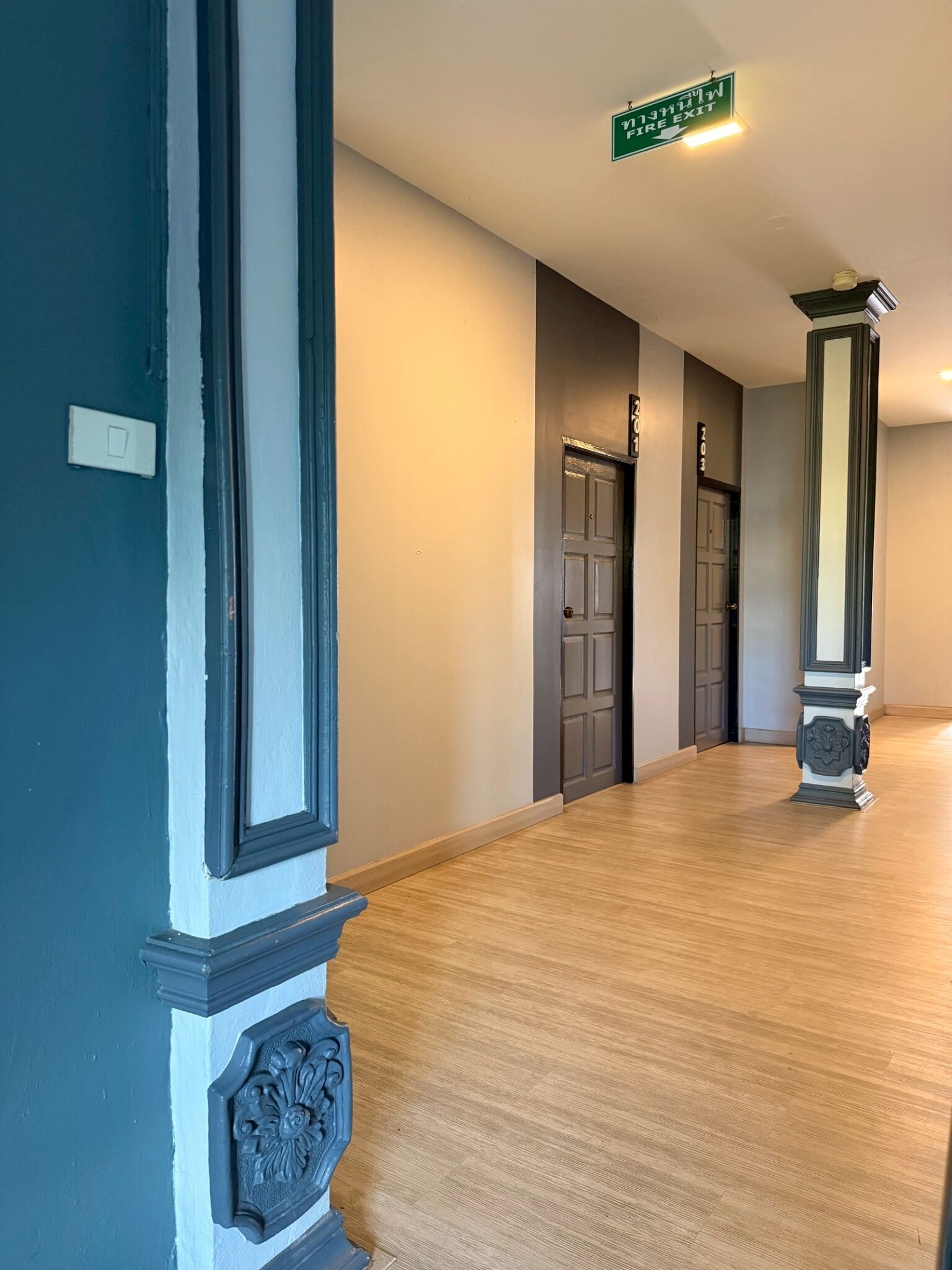 A hallway with a wooden floor, a green exit sign, and a door with a number above it.