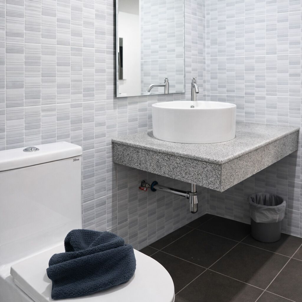 Un baño moderno con inodoro blanco, un lavabo redondo sobre encimera de granito, un espejo sobre el lavabo y un cubo de basura.