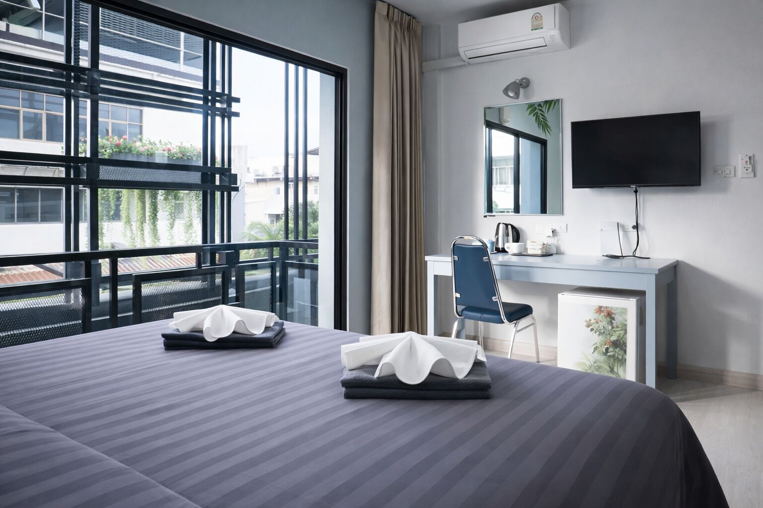 A hotel room with a large bed, a desk, a chair, a flat-screen TV, and a balcony with a view of buildings and greenery.