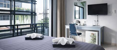 A hotel room with a large bed, a desk, a chair, a flat-screen TV, and a balcony with a view of buildings and greenery.
