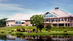 A large, modern building with a prominent roof and large windows, surrounded by a well-maintained lawn and a body of water. - Uniland Golf & Resort (Nakhon Pathom)