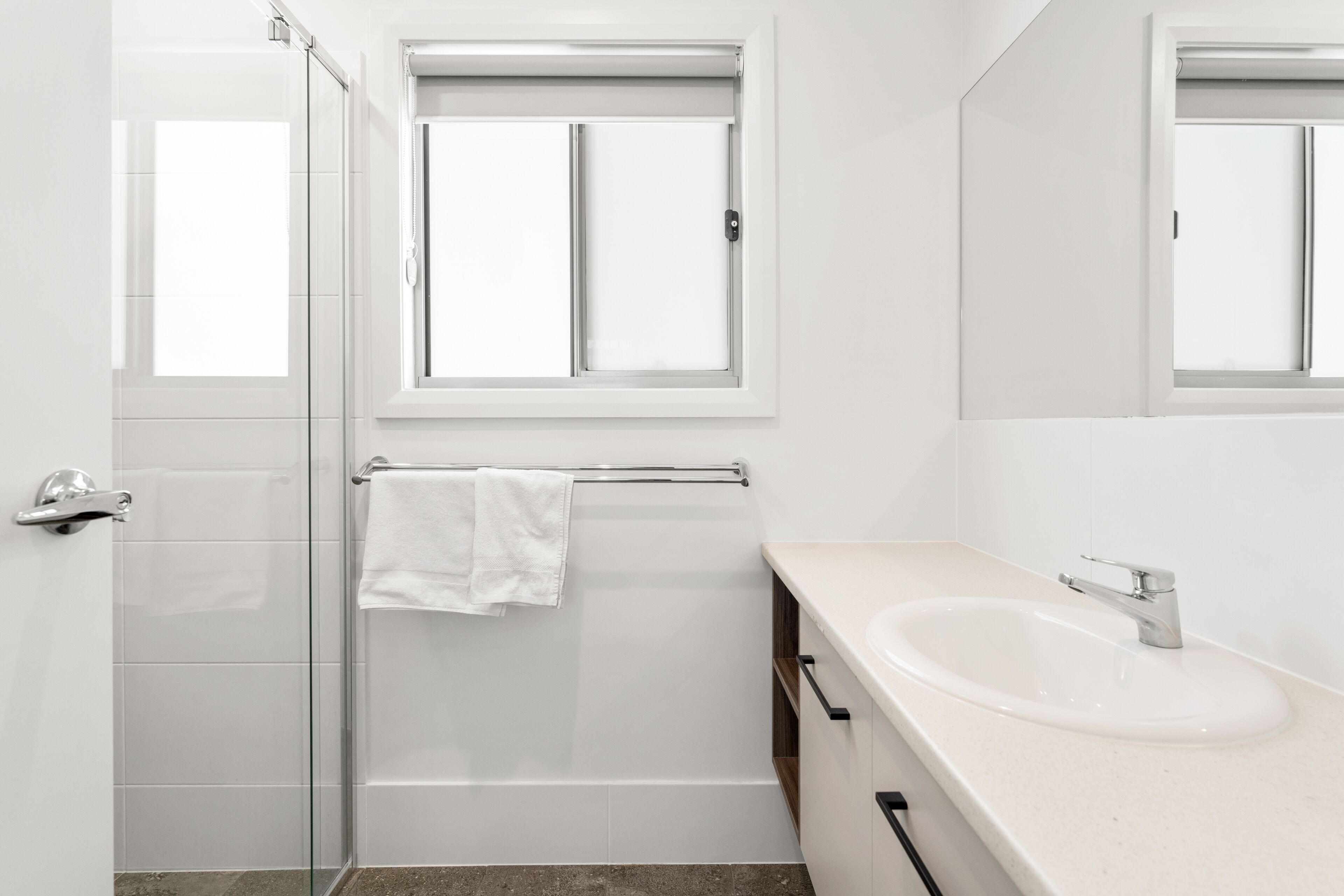 Un bagno moderno con cabina doccia in vetro, lavandino bianco e finestra sopra lo specchio.