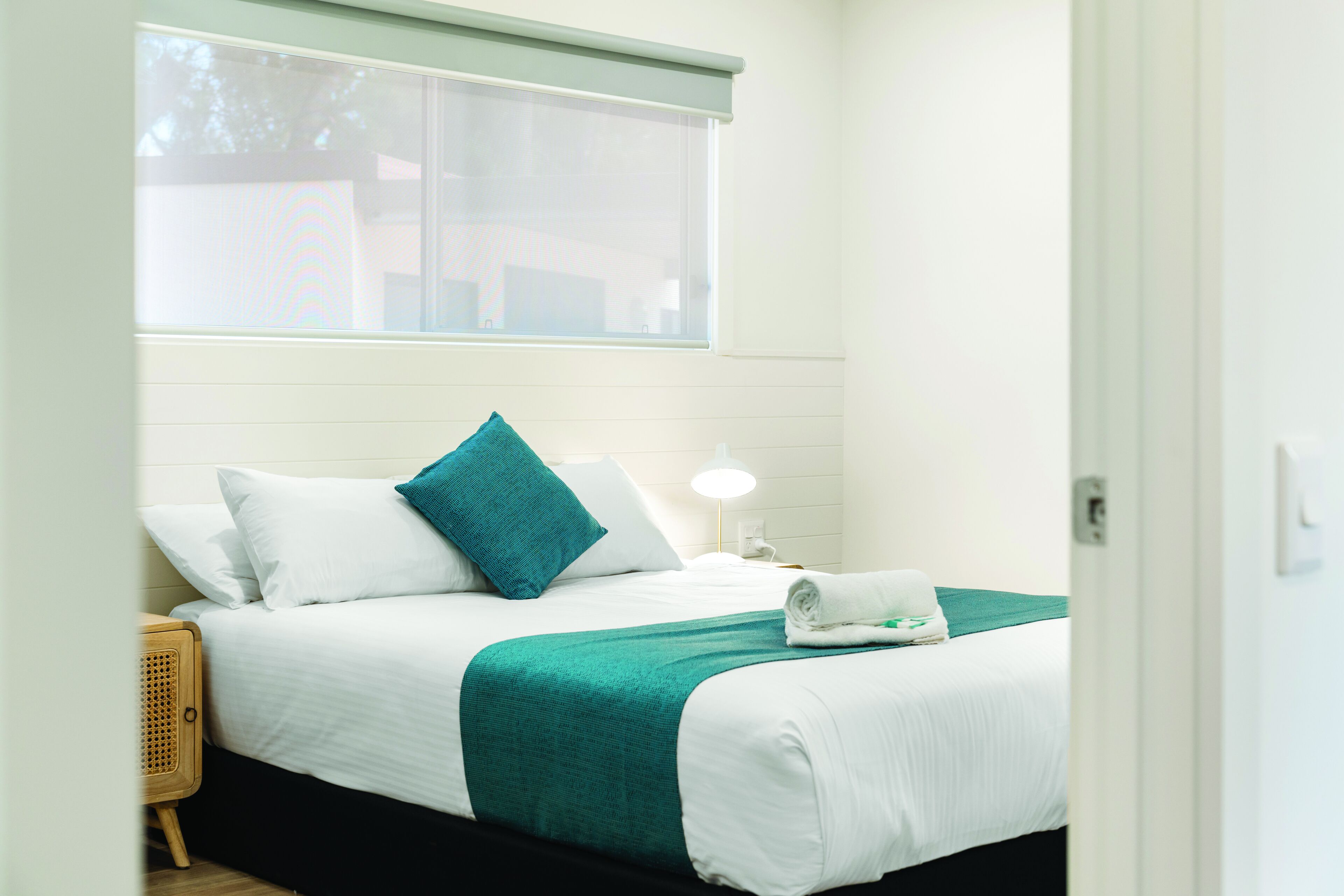 A neatly made bed with white and teal bedding, a bedside lamp, and a window with blinds.