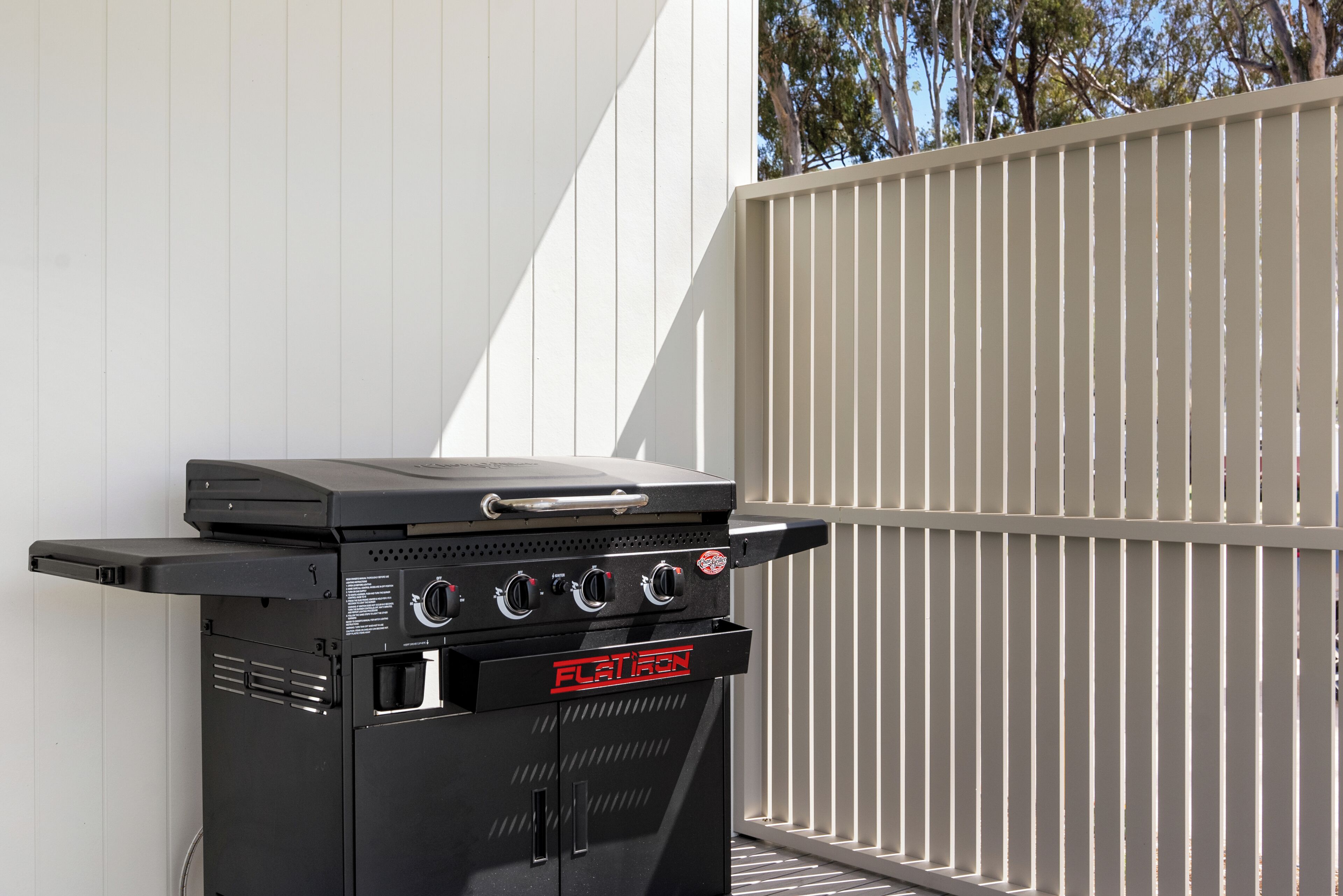 A black outdoor grill with a digital display and a sliding menu, positioned against a white wall with a metal fence in the background.