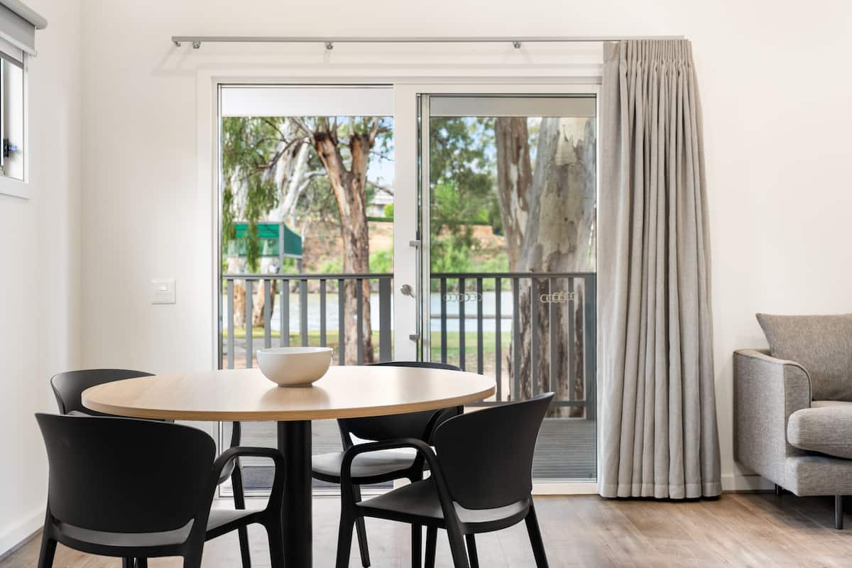 A dining area with a round table and four chairs, a sofa, and a sliding glass door leading to an outdoor area with trees and a fence.