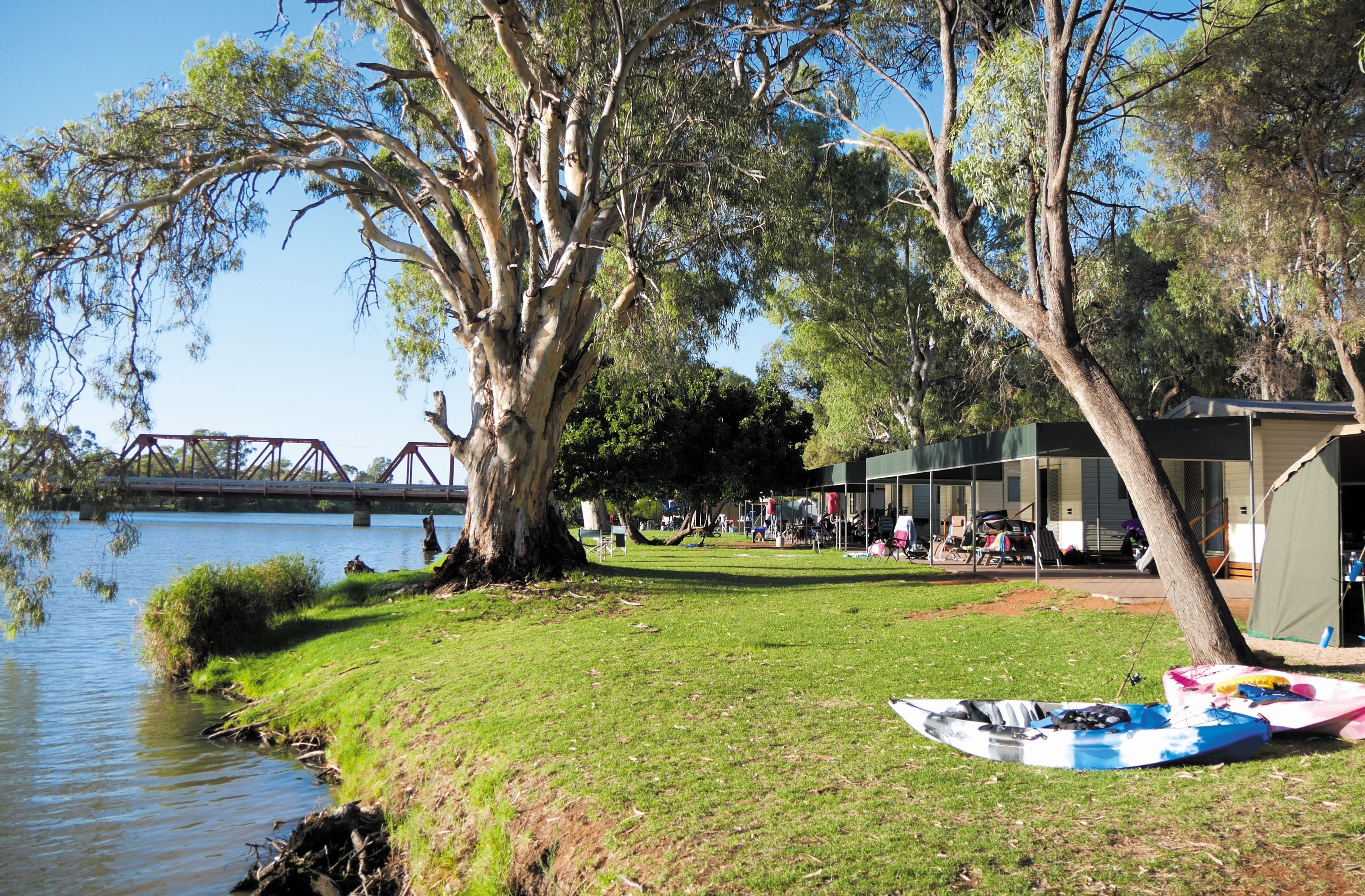 A riverside campsite with kayaks, a bridge in the background, and a building with outdoor seating.