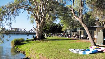 A riverside campsite with kayaks, a bridge in the background, and a building with outdoor seating.