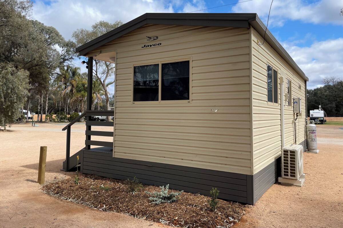 A small, single-story mobile home with the brand name 'Jayco' on the front. It has a small porch with a wooden bench and a single window on the side.