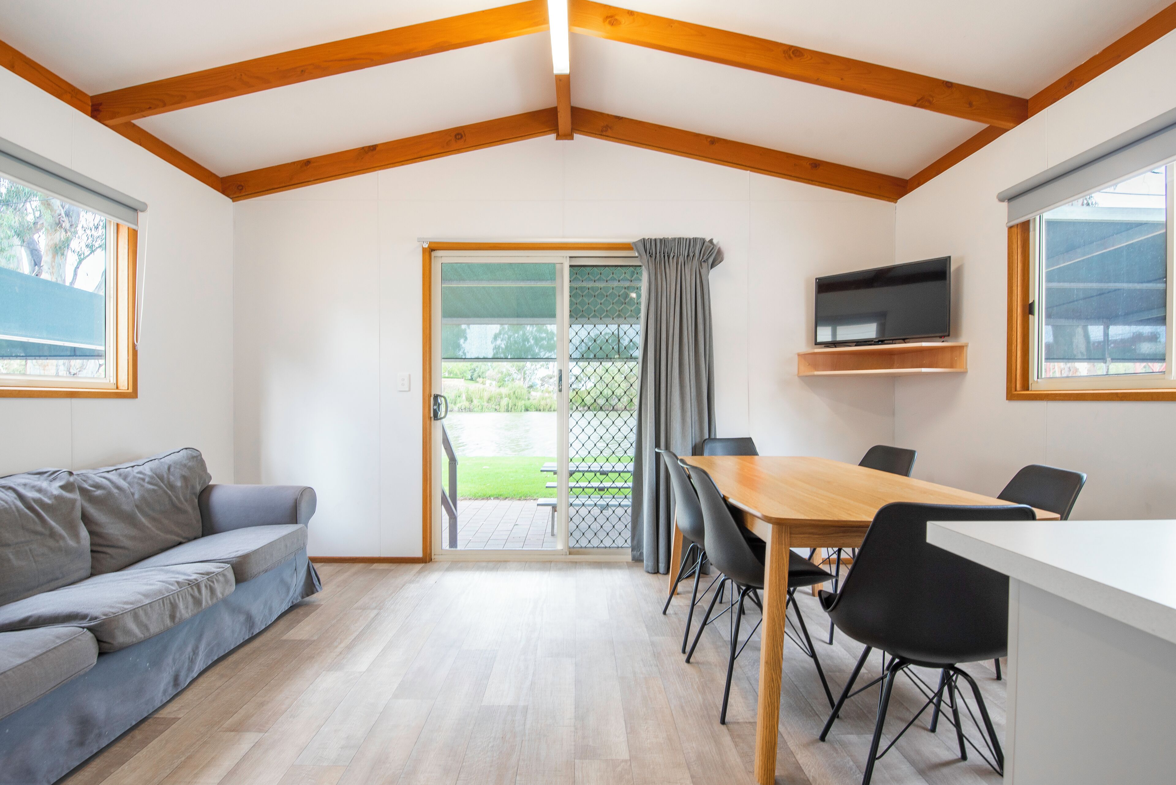 A spacious room with a wooden ceiling, a sofa, a dining table with chairs, a TV mounted on the wall, and a sliding glass door leading to an outdoor area.