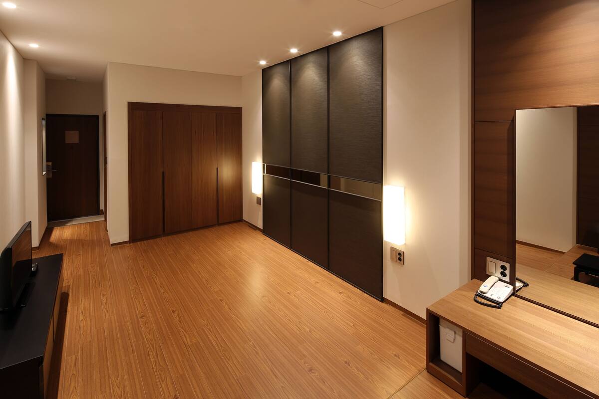 A modern hotel room with a wooden floor, a large wardrobe, a bedside table with a phone, and wall-mounted lighting.