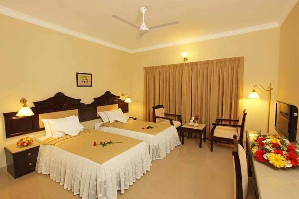 A hotel room with two beds, a ceiling fan, a TV, a desk with a lamp, and a chair.
