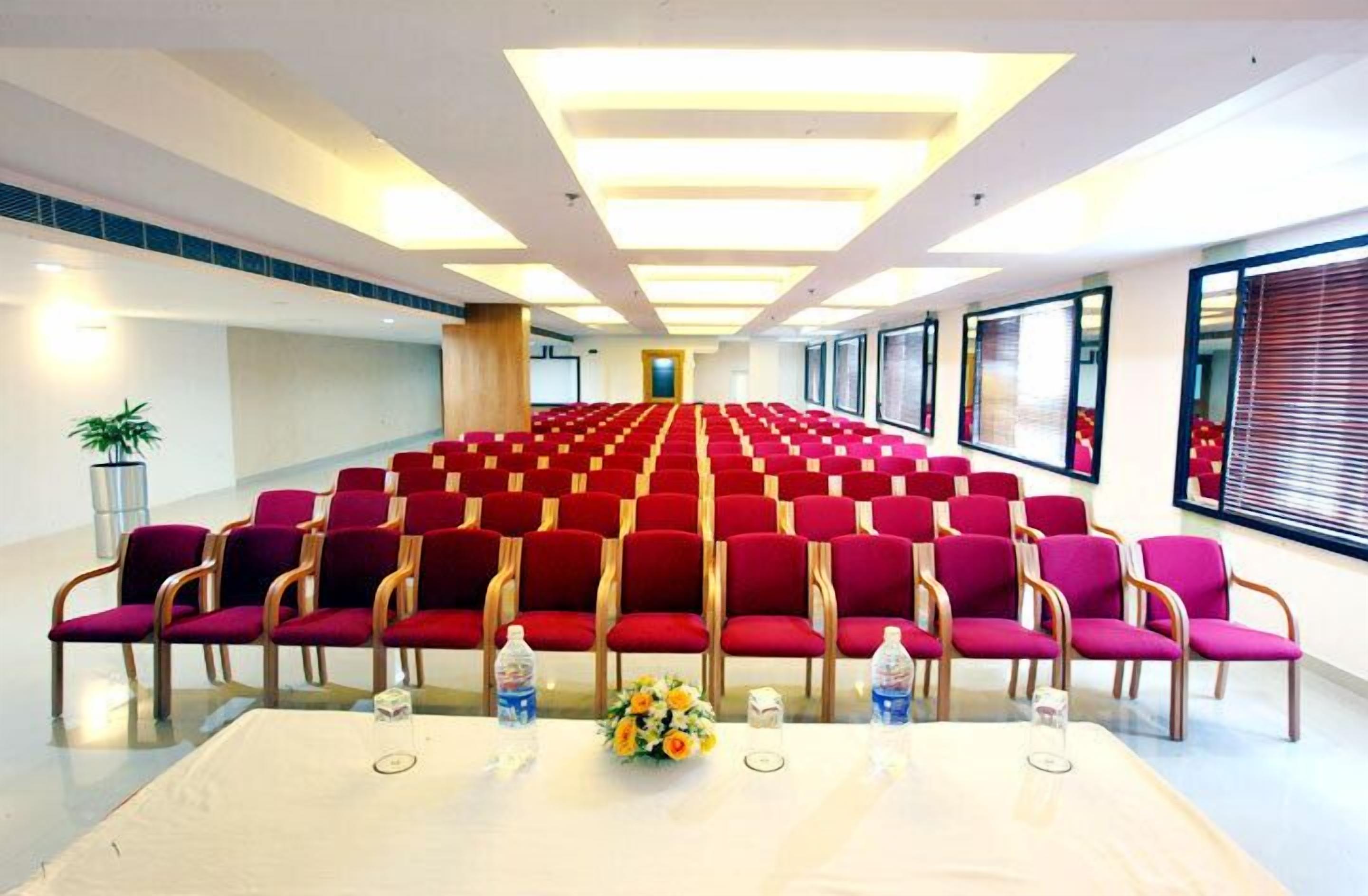 Un salón de conferencias con filas de sillas rojas, una larga mesa con botellas de agua y un arreglo floral, y amplias ventanas con persianas.
