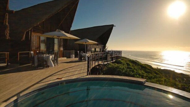 Ein Poolbereich mit Blick auf das Meer bei Sonnenuntergang, mit Sitzgelegenheiten im Freien und einer Holzterrasse.