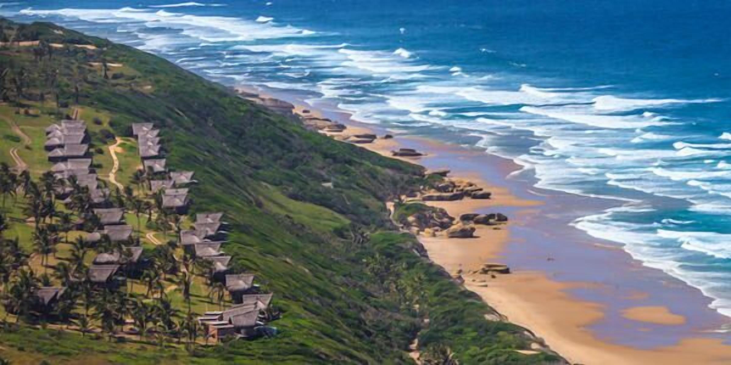A coastal resort with villas on a hillside, a sandy beach, and the ocean.
