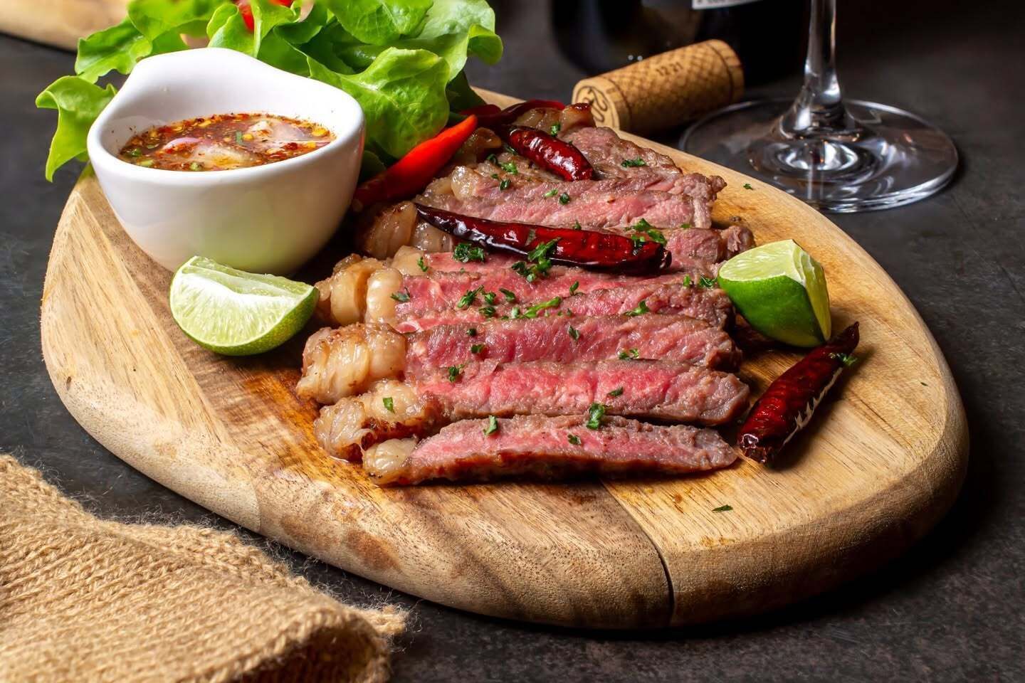 A wooden cutting board with sliced steak, lime wedges, and chili peppers.