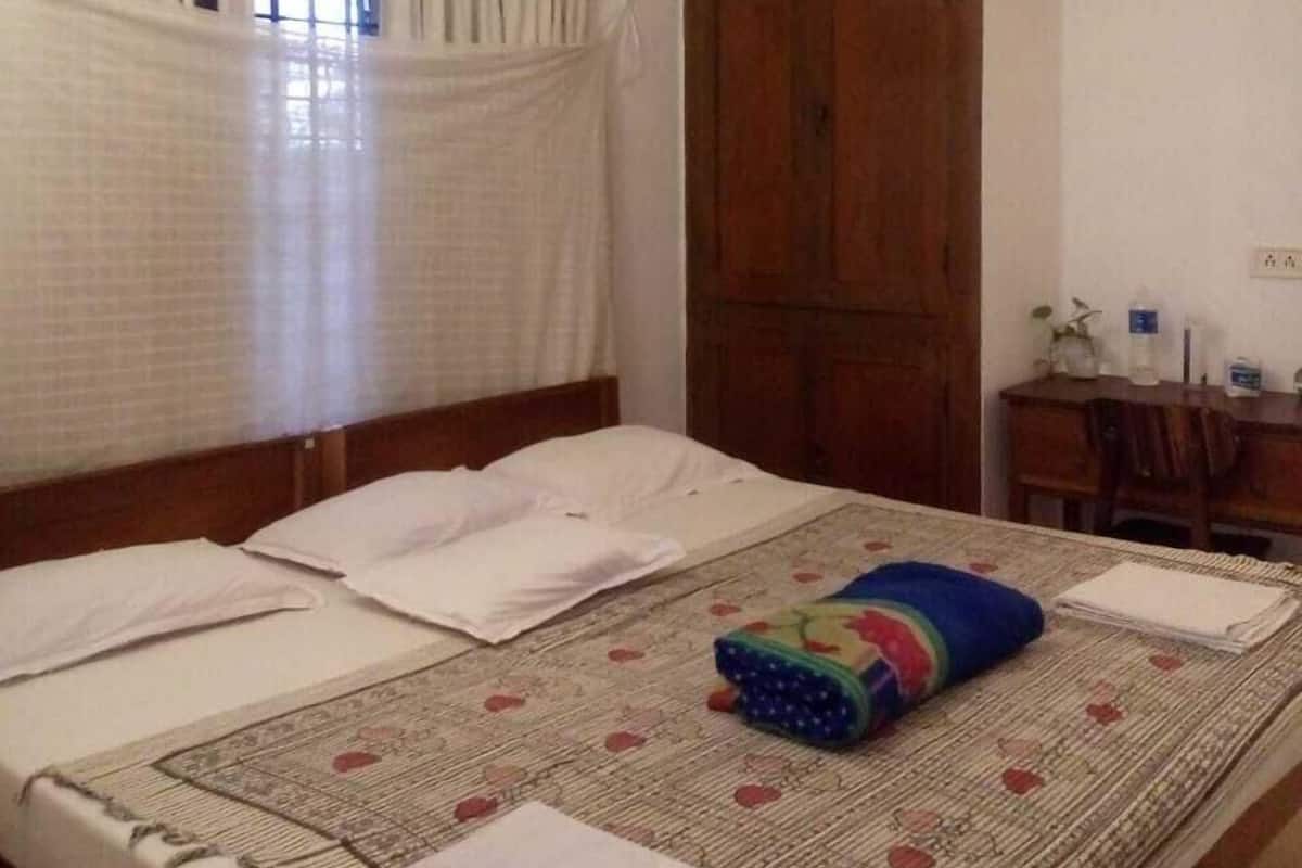 A bedroom with a bed, a ceiling fan, an air conditioning unit, a window with curtains, a wooden door, and a small desk with a chair.
