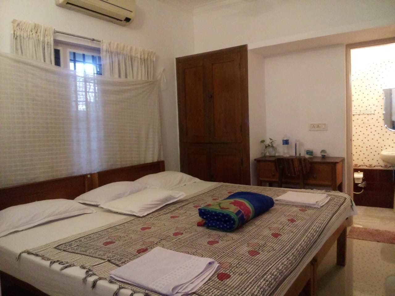 A bedroom with two beds, a wooden wardrobe, a desk with a chair, and a bathroom visible through an open door.