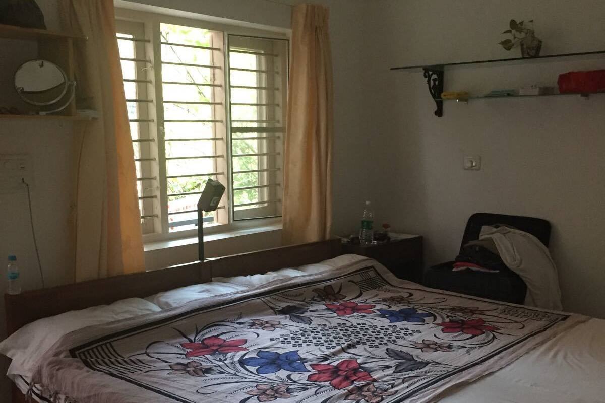 A bedroom with a bed, a window with curtains, an air conditioning unit, and a shelf with items.