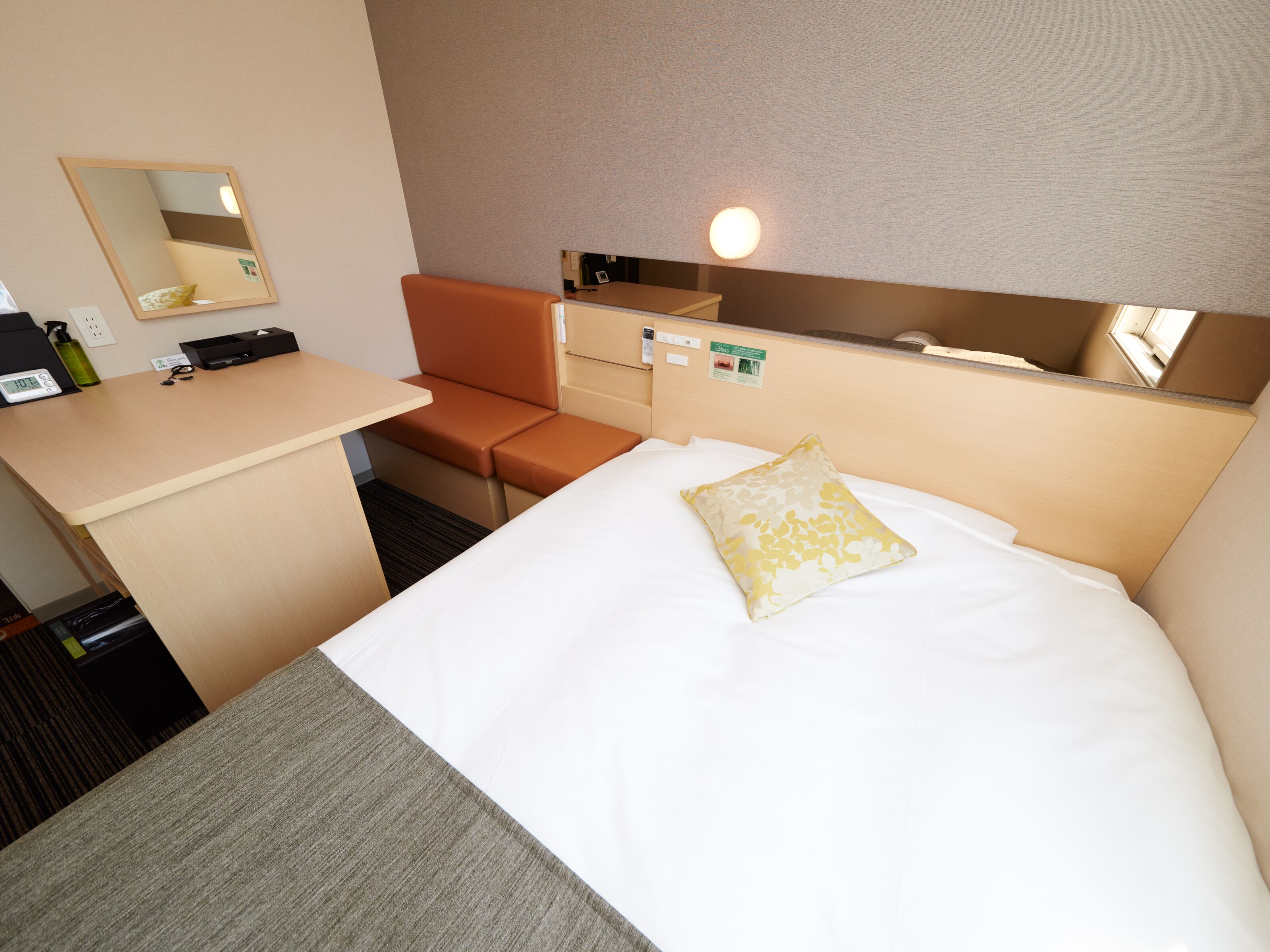 Habitación de hotel con cama, escritorio, silla, mesita de noche, espejo y lámpara de pared.