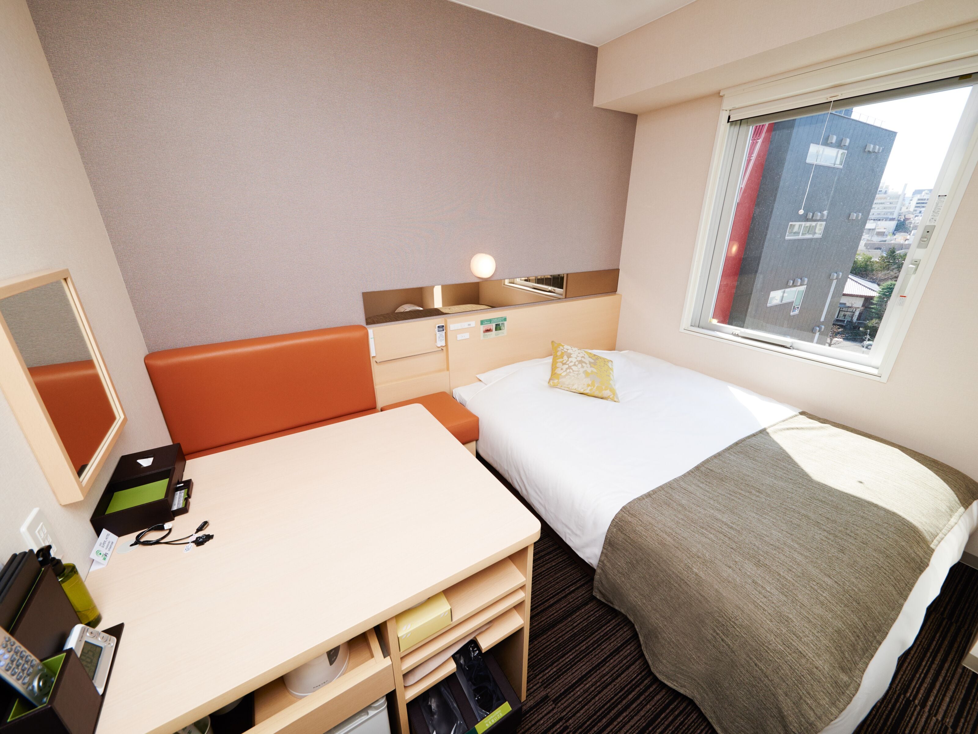 A hotel room with a bed, a desk, a chair, a mirror, a window, and a view of buildings outside.