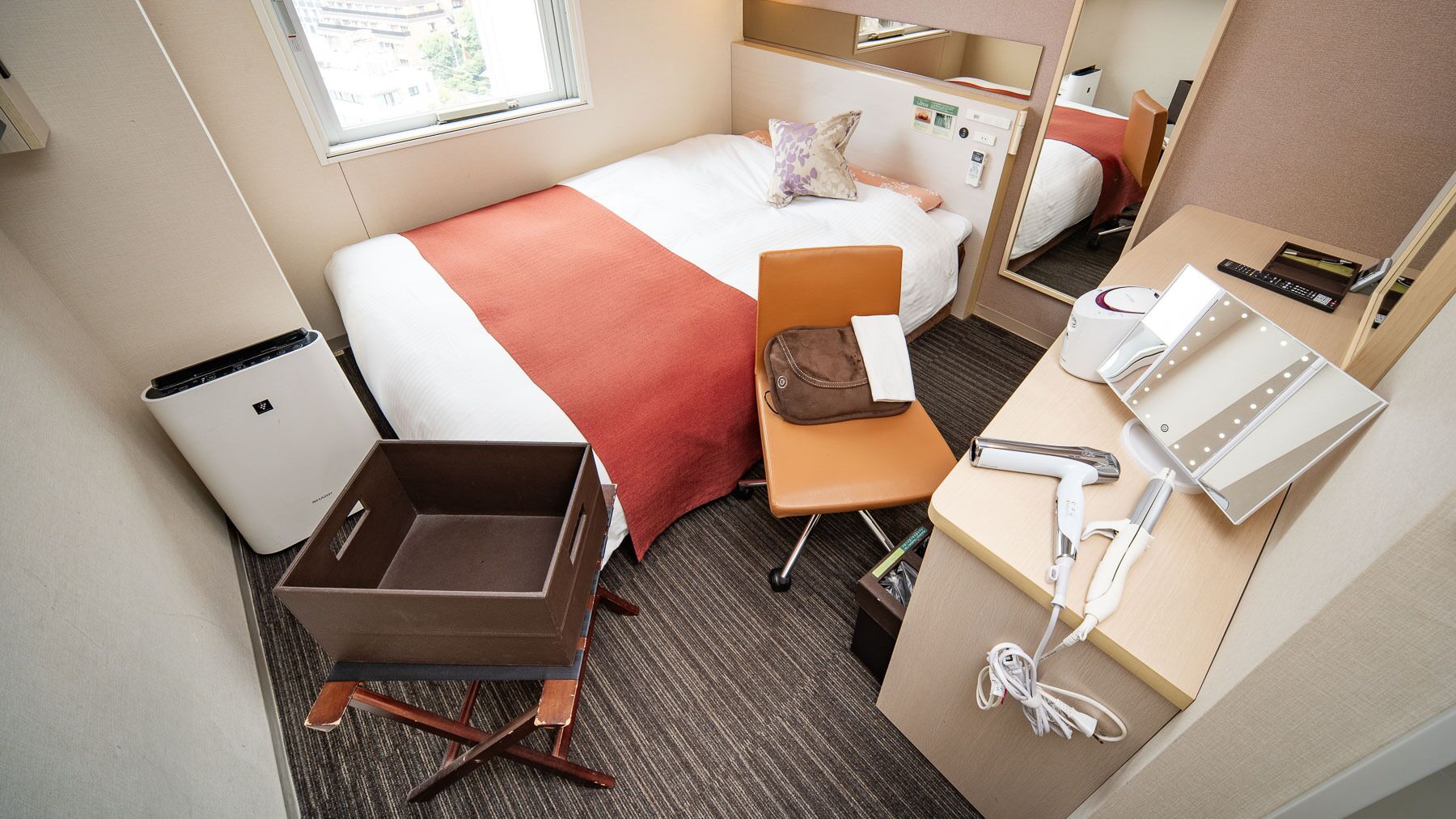A hotel room with a bed, a desk with a mirror, a small trash bin, and a suitcase.