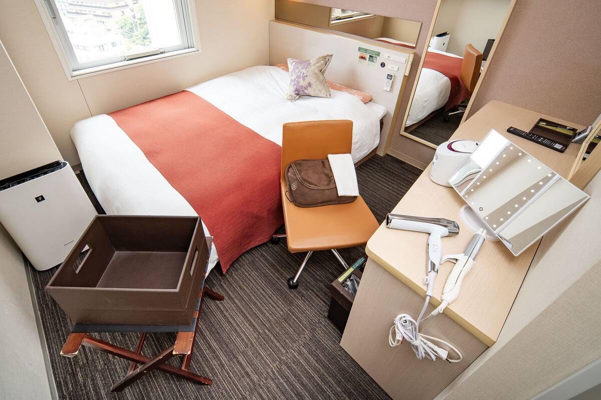 Une chambre d’hôtel avec un lit, un bureau doté d’un miroir, une petite poubelle et une valise.